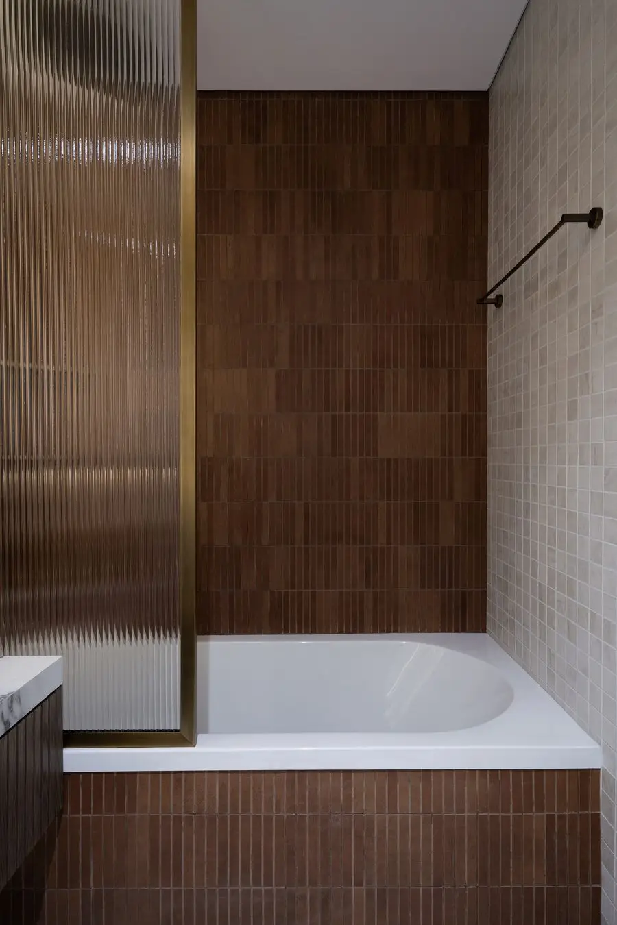 Bathtub framed by vertical copper-toned tile and a fluted glass screen